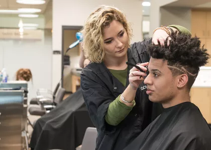 A barbering student shaving the side of a young man's head.