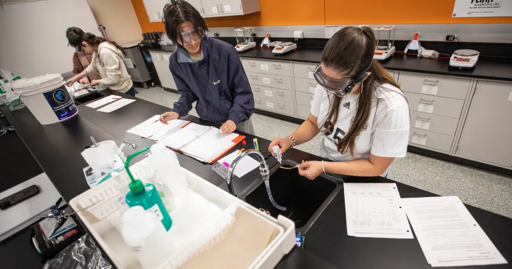 Two students in a lab working on a project, wearing eye googles and putting solution in an eye dropper on a slide.