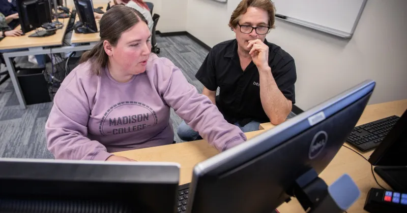 Student, wearing purple sweatshirt, working on desktop computer with instructor by her side.
