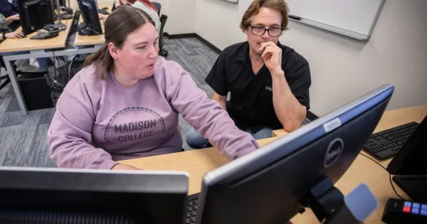 Student, wearing purple sweatshirt, working on desktop computer with instructor by her side.