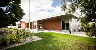 Image of the outside view of the Watertown campus showing people walking in the door. Landscape of green grass and trees.