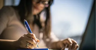 Woman with brown hair holding pen and writing in a notebook on a desk.