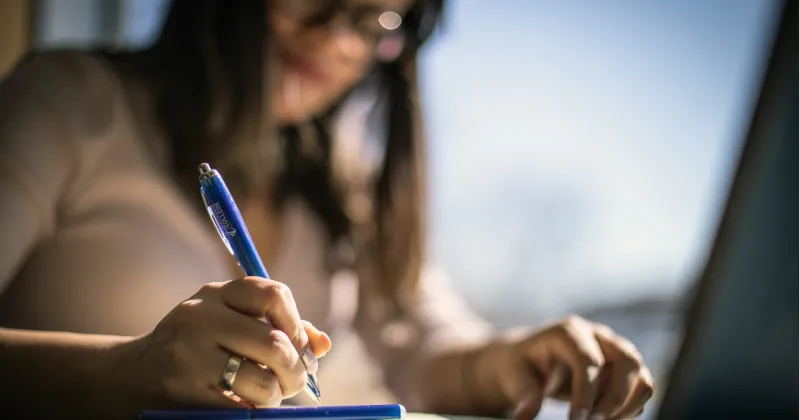 Woman with brown hair holding pen and writing in a notebook on a desk.