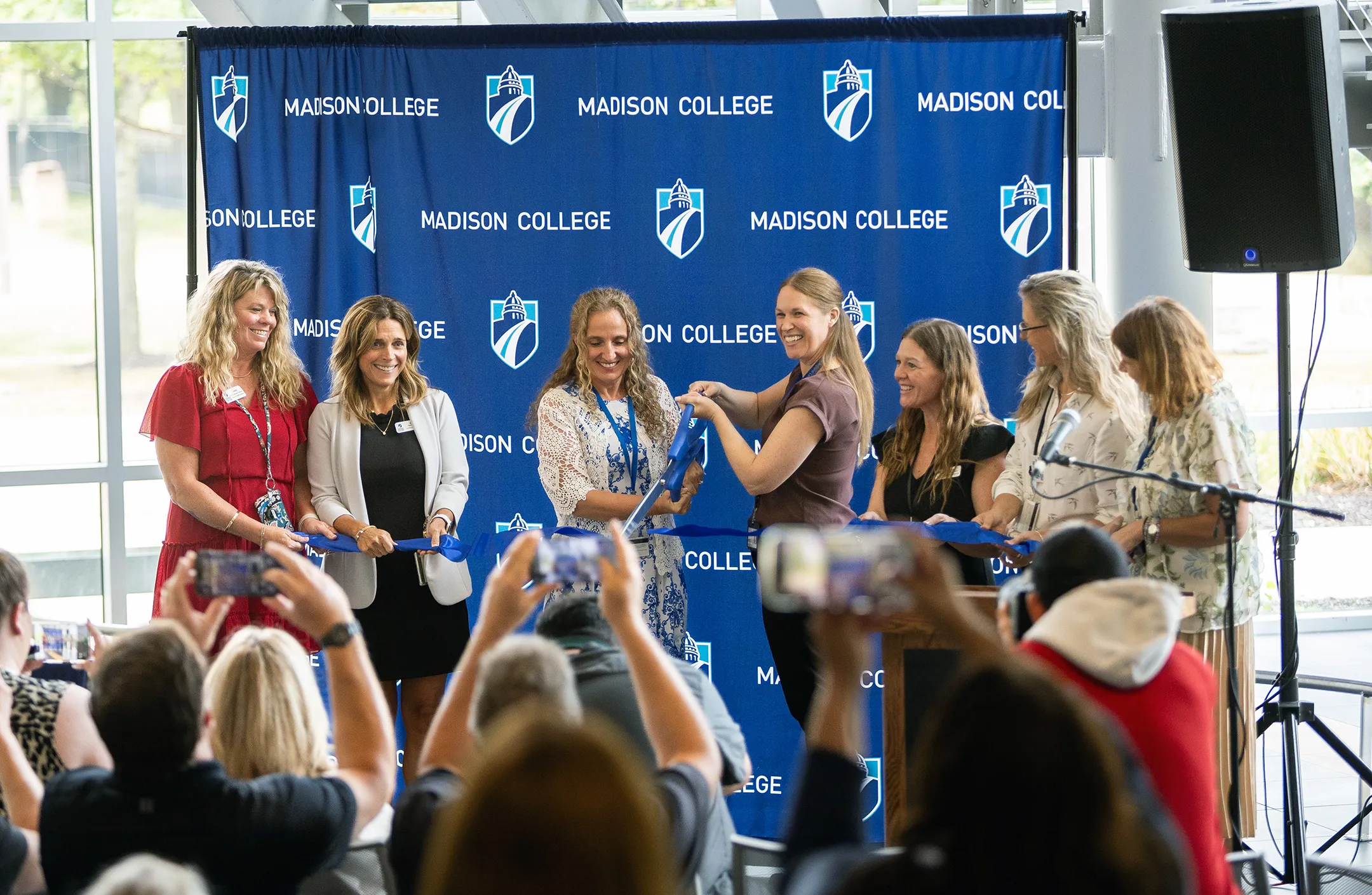 Staff and faculty standing in front of a blue banner with one person cutting a ribbon during a ceremony.