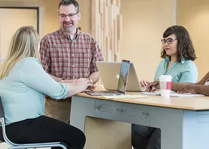 Teacher is standing and talking to two students seated at a table.