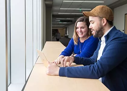 Two happy students seated at a long table facing a window looking at a tablet.