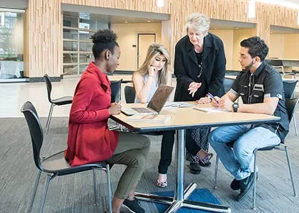 Teacher is standing and talking to three students seated at a table.