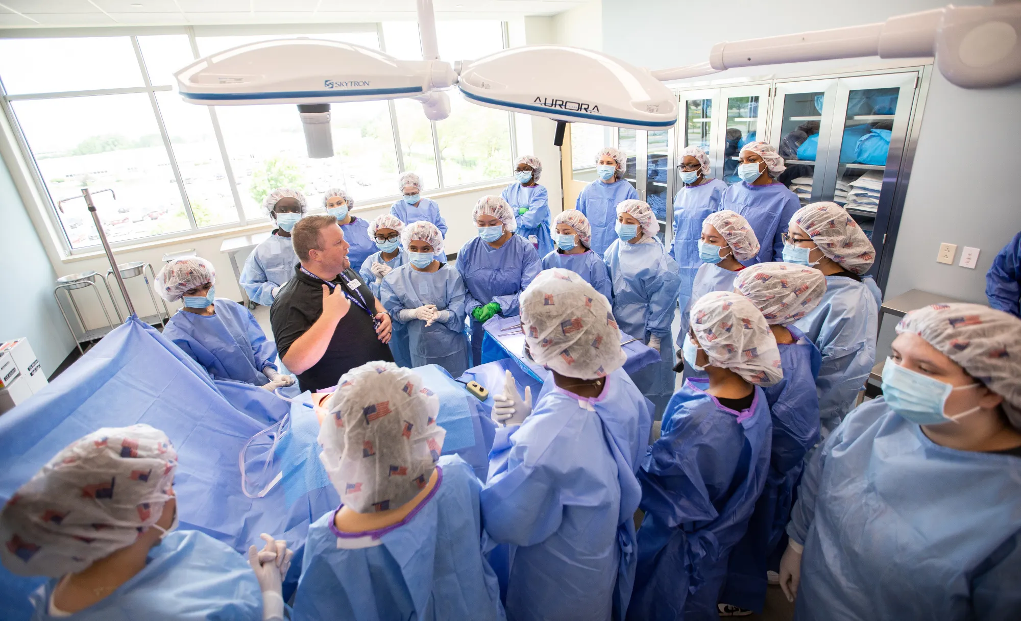 Surgical technology students in scrubs gathered around an instructor in a simulated operating room at the Truax campus.