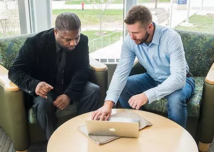 Two students seated at a round table looking at a macbook.