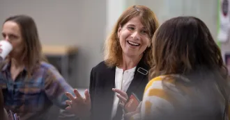President Jennifer Berne, wearing dark blazer, talking with a person in a yellow striped sweater.
