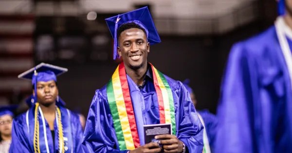 Black graduate,in blue cap and gown and African color shawl, walking in a commencement ceremony.