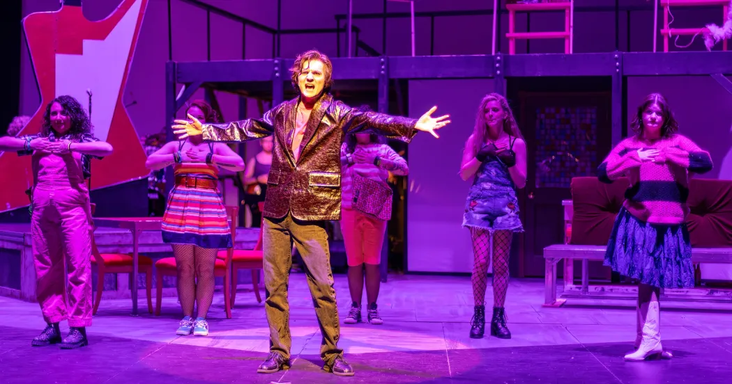 Student in brown coat and pants on a stage singing with arms outstretched.