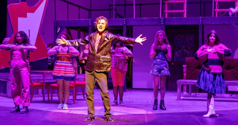 Student in brown coat and pants on a stage singing with arms outstretched.