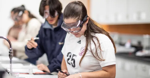 Student in a lab setting wearing protective eye gear and looking at a notebook with a pen in her hand.