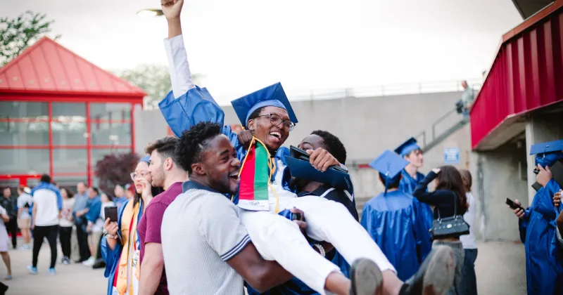 Black male graduate, dressed in blue cap and gown, being held up in the air, by two friends in celebration of his graduation.