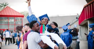 Black male graduate, dressed in blue cap and gown, being held up in the air, by two friends in celebration of his graduation.