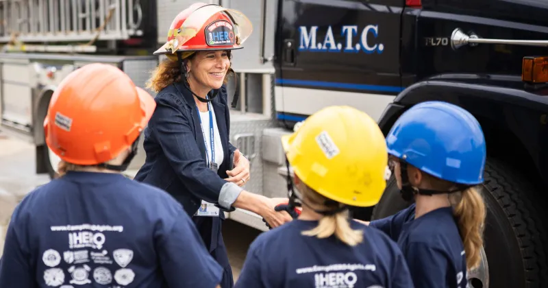 President Berne, wearing a firefighter helmet, shakes hands with young girls in the CampHero program.