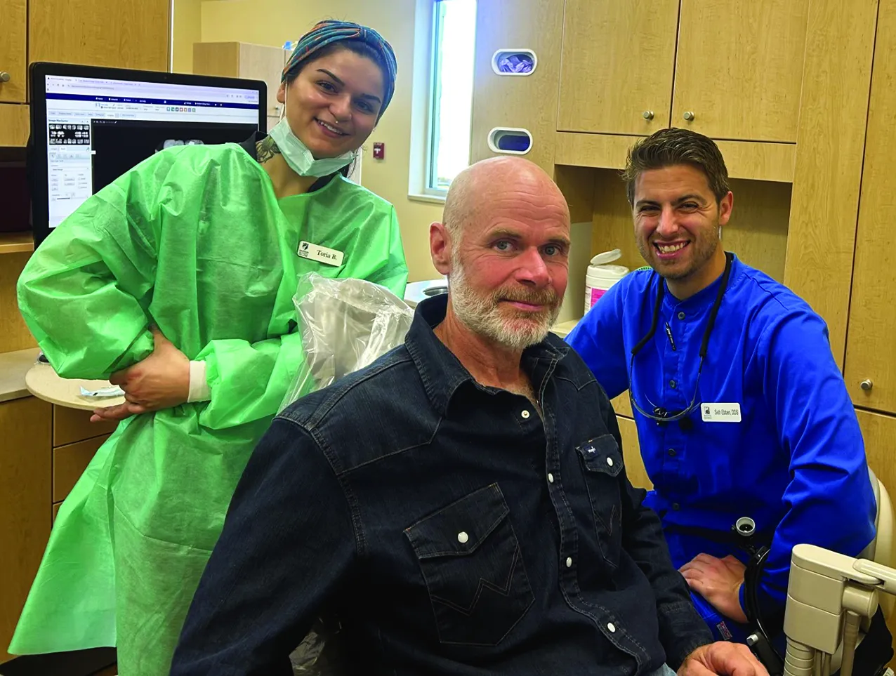 Dental patient sitting in a clinic chair, while a dental hygienist and dentist look on, smiling.